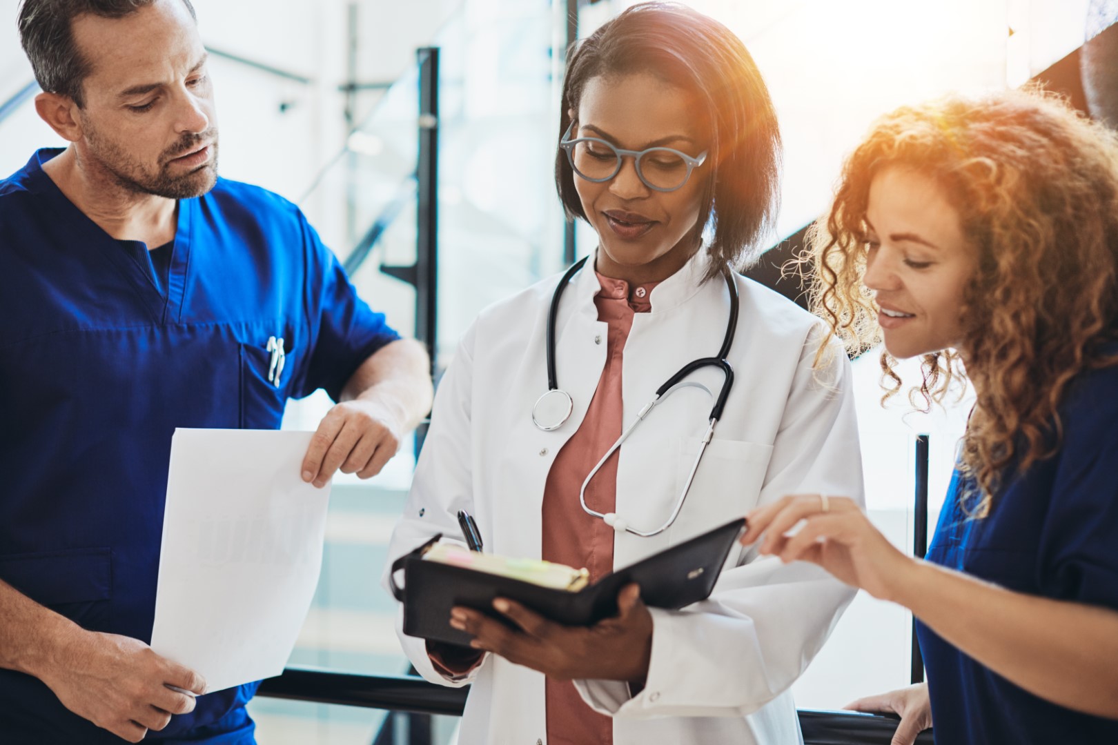 Healthcare professionals in scrubs and a white coat reviewing notes and documents together in a clinical setting.