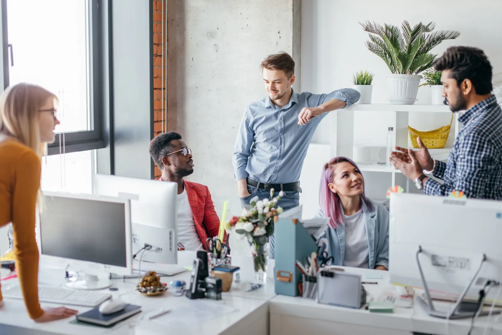 Small business team having an informal discussion around desks in a bright office with computers and decorative plants.