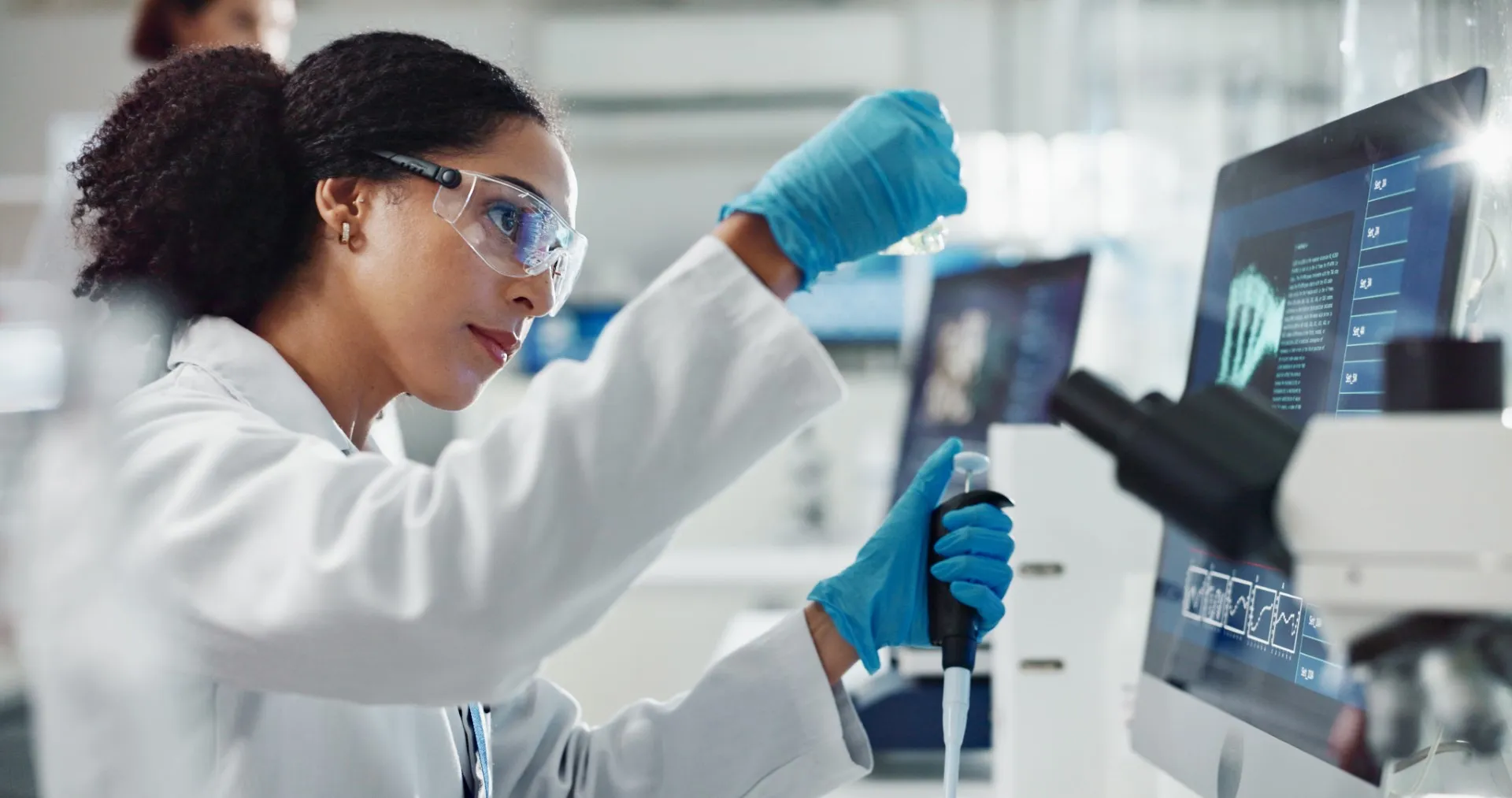 Scientist in a laboratory using a pipette while analyzing DNA data displayed on a computer screen.