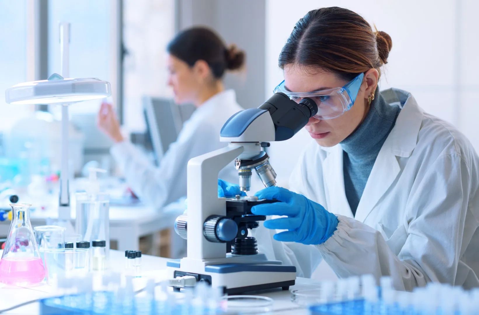 Scientist in a laboratory examining samples through a microscope with test tubes and lab equipment on the table.