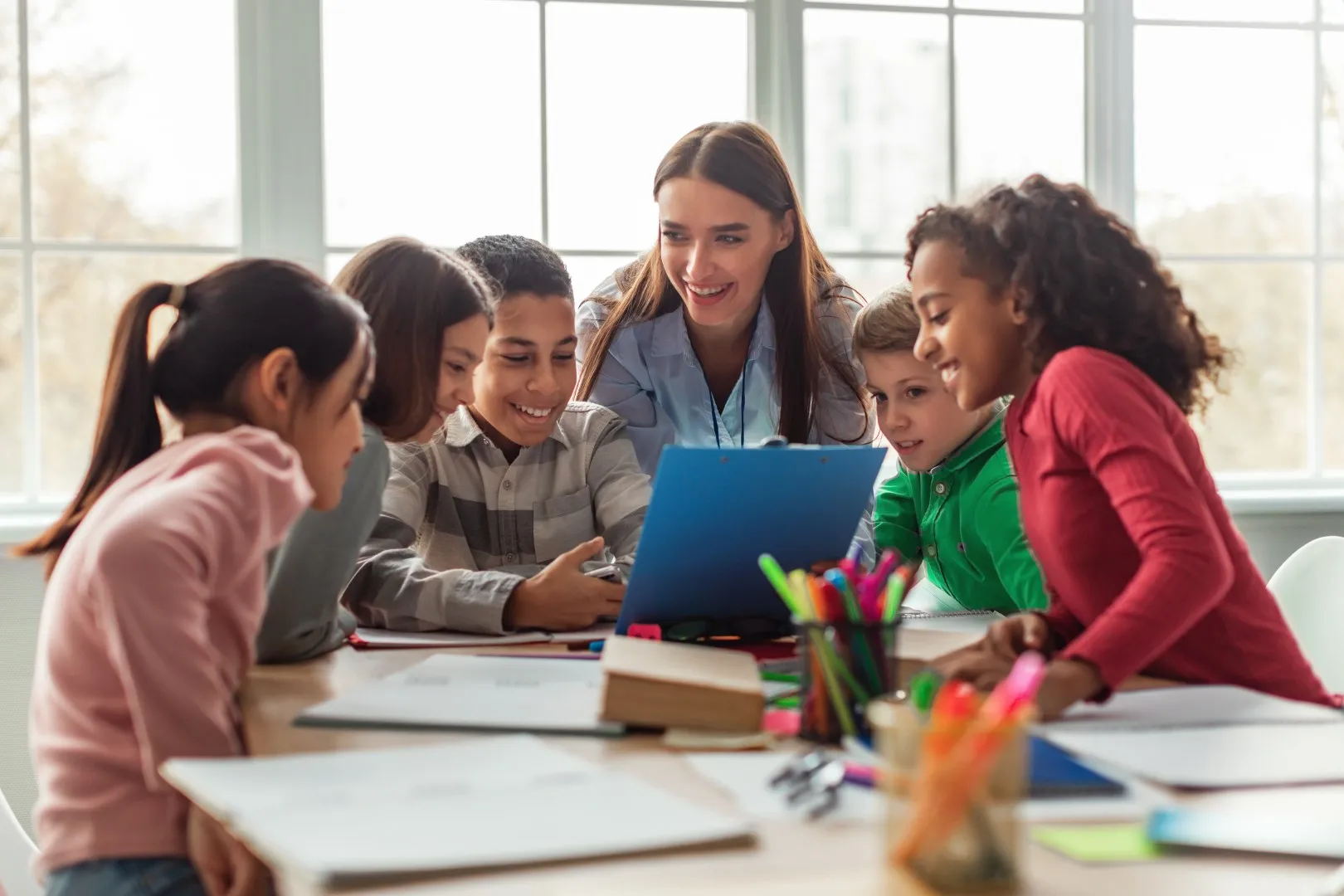 Teacher guiding a group of students gathered around at a classroom table with notebooks and colorful stationery.