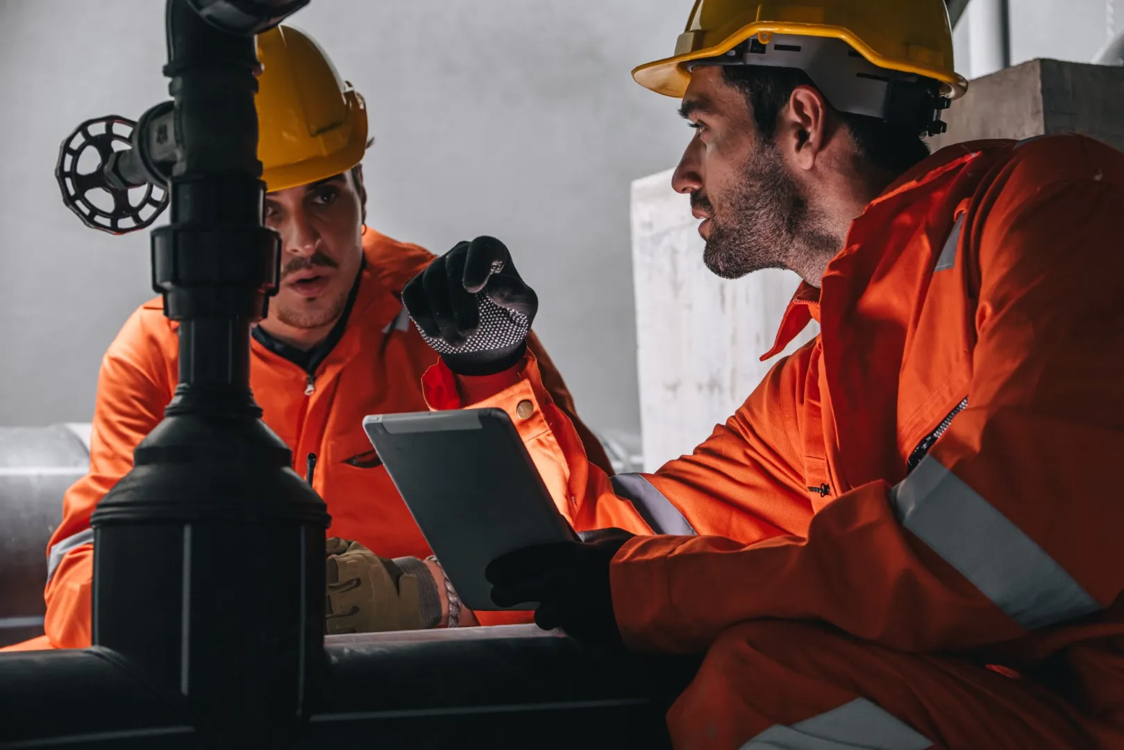 Utility workers wearing safety helmets and orange protective clothing inspecting equipment and reviewing information on a tablet inside an industrial facility.