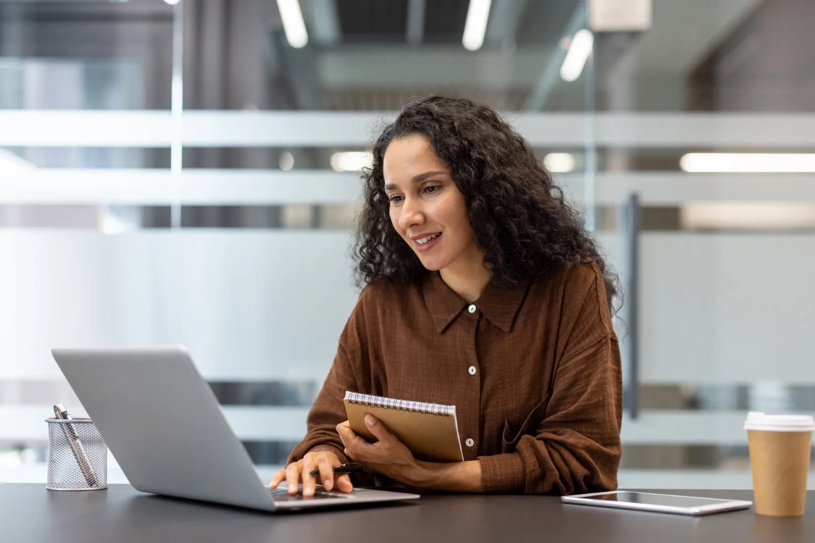 Employee working at a laptop while holding a notebook in a modern office, reflecting focus, initiative, and thoughtful problem‑solving